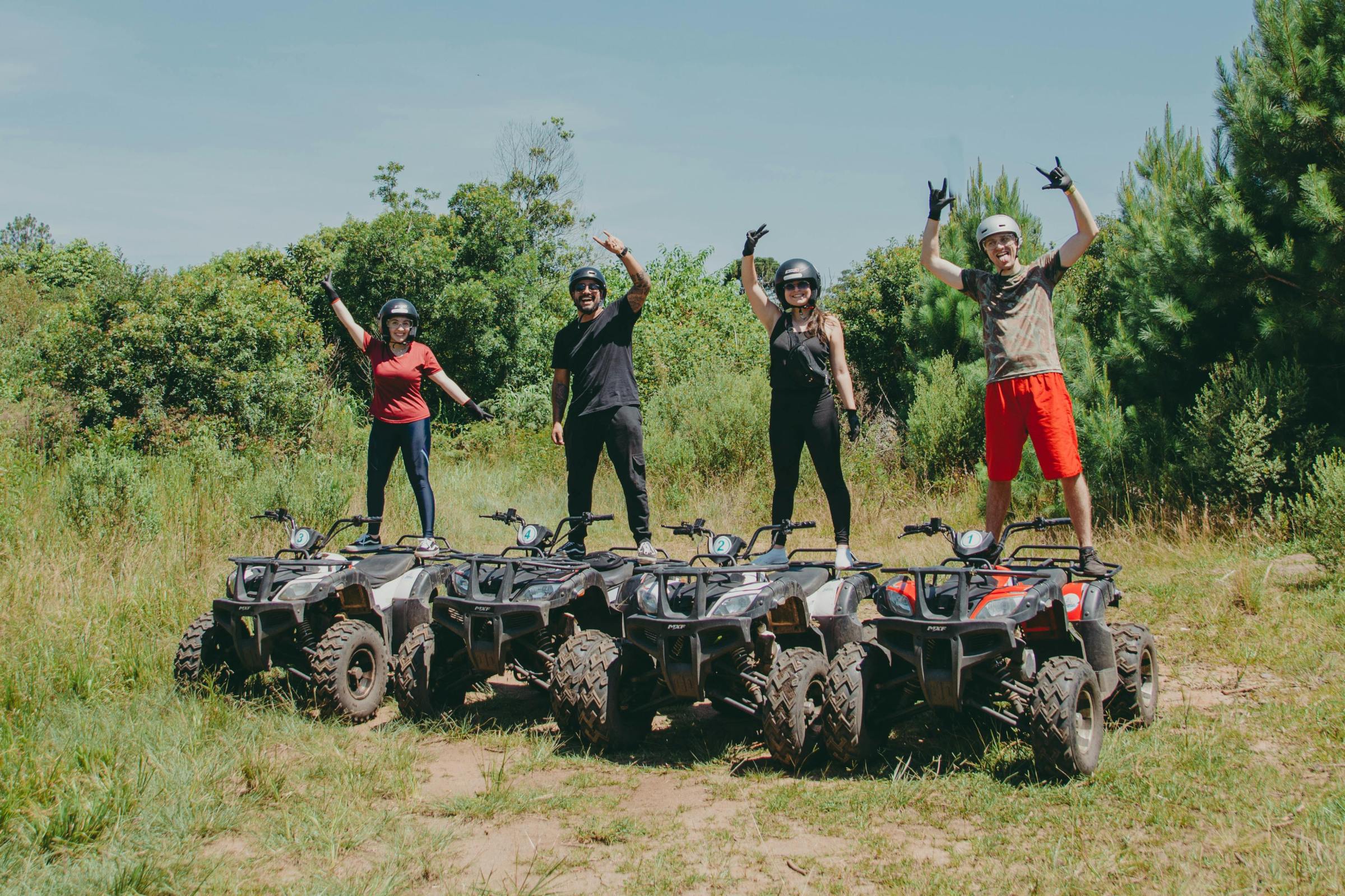 Four people stand on ATVs in a field with trees, raising arms in excitement.